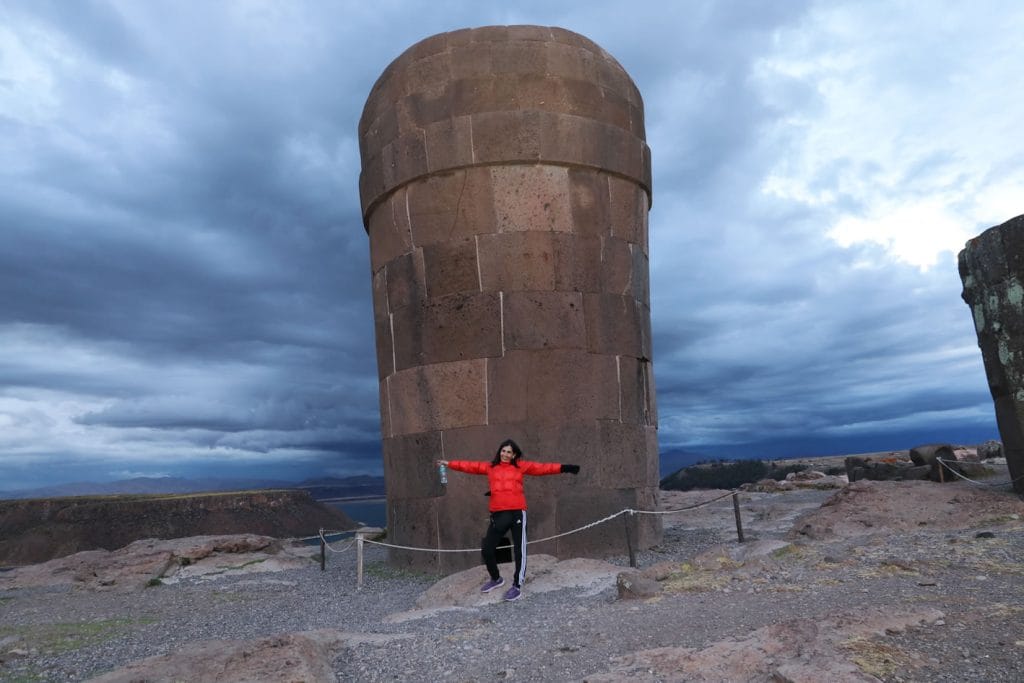 Mujer posando frente a una chullpa de la cultura Ccolla en el complejo arqueológico de Sillustani, con nubes oscuras en el cielo de la laguna Uyumayo.