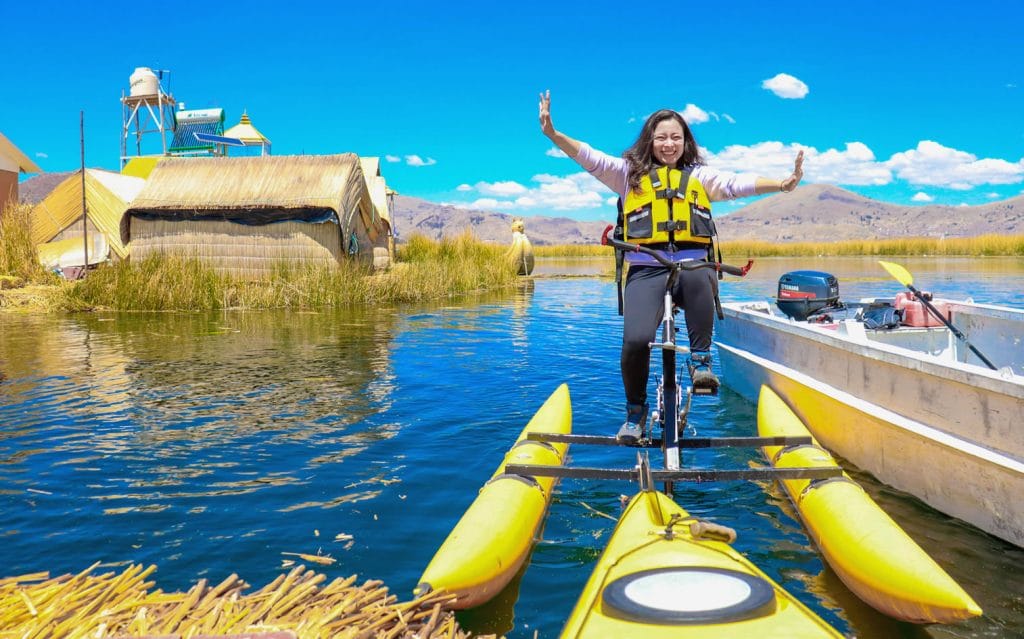 Mujer turista sonriente disfrutando del kayak, detrás una casa flotante de los Uros y el totoral visible.