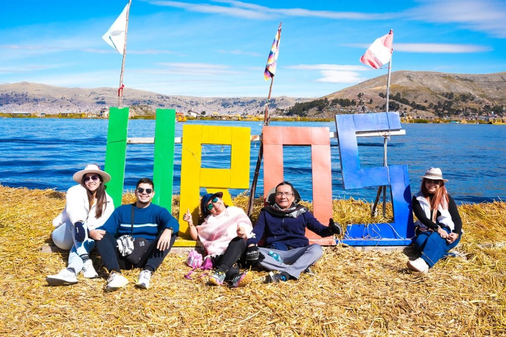 Grupo de turistas posa con letrero "UROS" de totora; vista panorámica de la ciudad de Puno al fondo, Lago Titicaca.