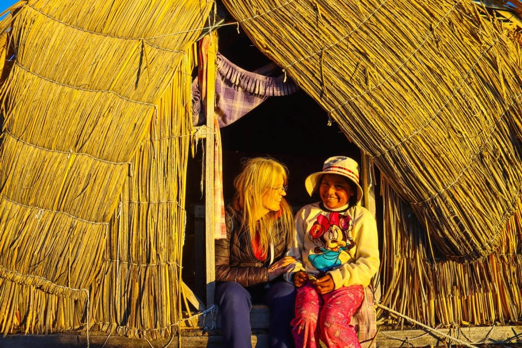 Felicidad y conexión: Niña Uros sonríe a cámara mientras turista la mira con calidez, sentadas en la puerta de choza de totora.