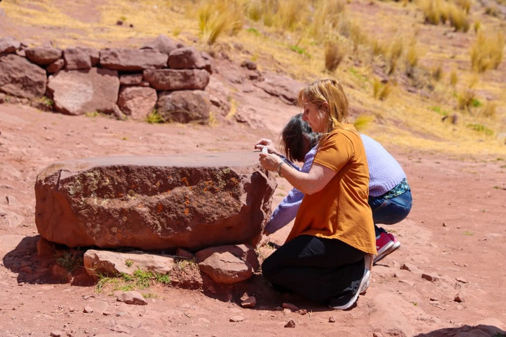 Dos mujeres turistas arrodilladas junto a la mesa ceremonial de piedra en el portal interdimensional de Aramu Muru (Hayu Marca), de camino a Tiahuanaco desde Puno-Perú.