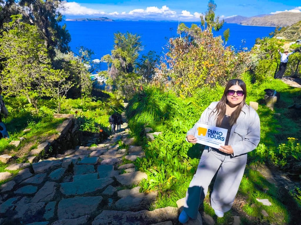 Turista sosteniendo el logo de Puno Tours al borde de un sendero rústico, durante el ascenso a un mirador o sitio arqueológico en el tour de la Isla del Sol.