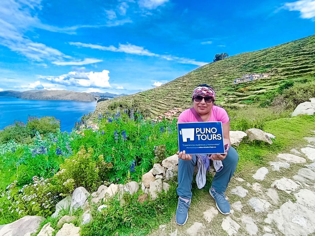 Mujer turista sentada en una calle de piedra inca de la Isla del Sol, sosteniendo el logo de Puno Tours con una vista espectacular del Lago Titicaca al fondo.