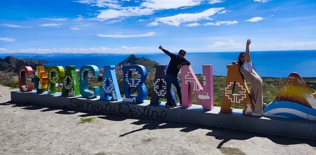 Pareja de turistas posando apoyados junto a las grandes letras monumentales que forman el nombre COPACABANA, con el Lago Titicaca al fondo, celebrando su llegada.