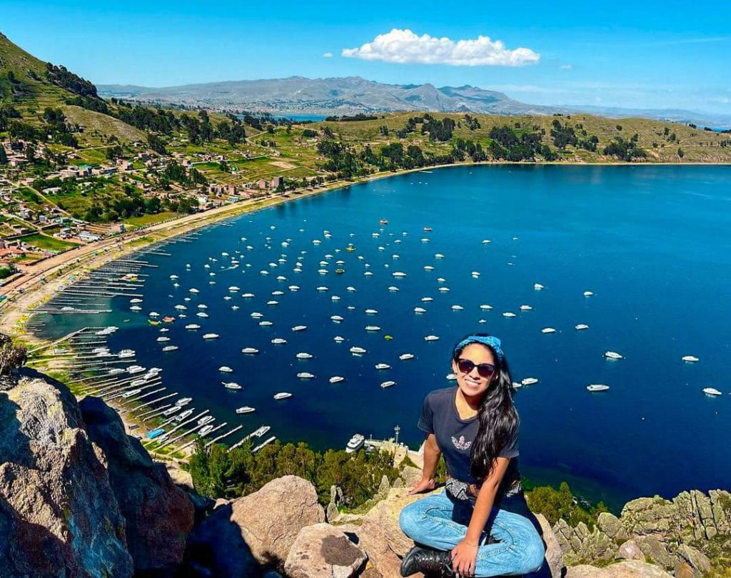 Mujer turista sonriente en primer plano, sentada y disfrutando de la vista panorámica de la bahía y la ciudad de Copacabana, a orillas del Lago Titicaca en Bolivia.