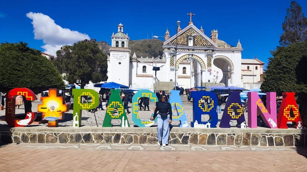 Mujer viajera posando junto a las grandes letras COPACABANA, con la icónica Basílica de Copacabana visible al fondo, en la orilla del Lago Titicaca, Bolivia.