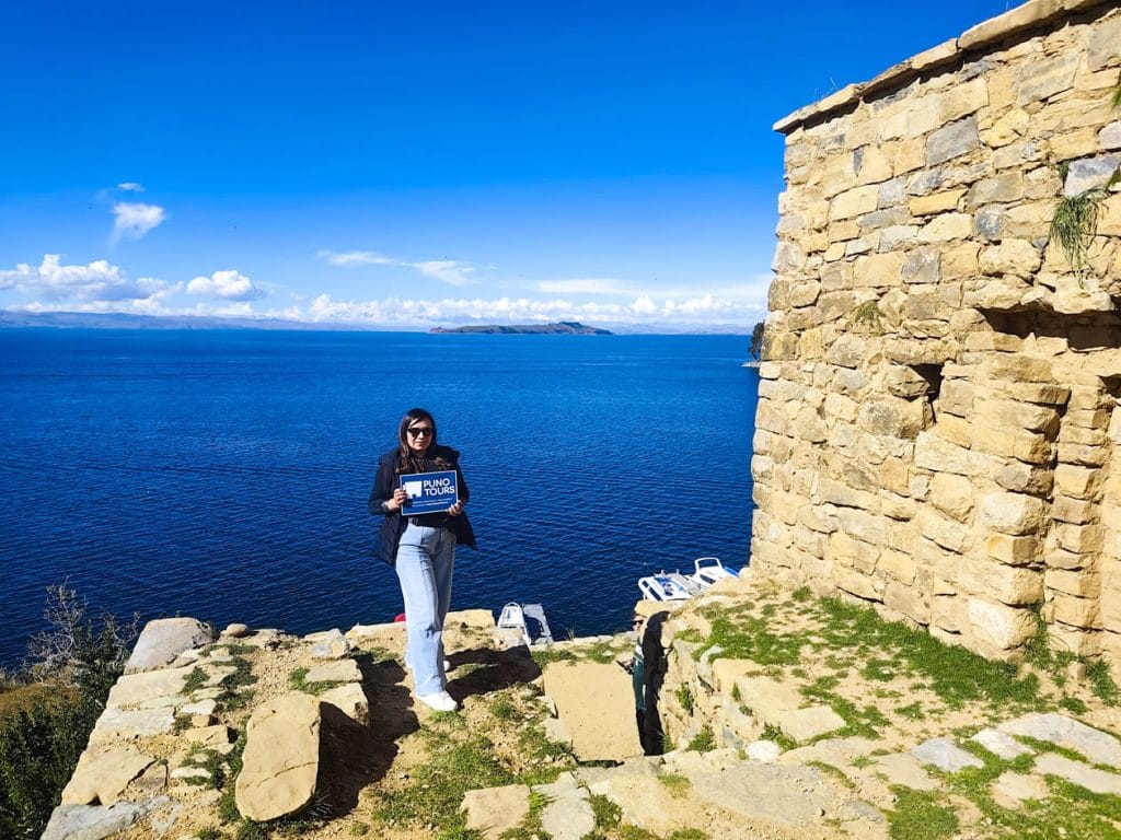 Mujer turista de pie sosteniendo el logo de Puno Tours, posando junto a las antiguas paredes de piedra de un templo ancestral inca en la Isla del Sol, Bolivia.
