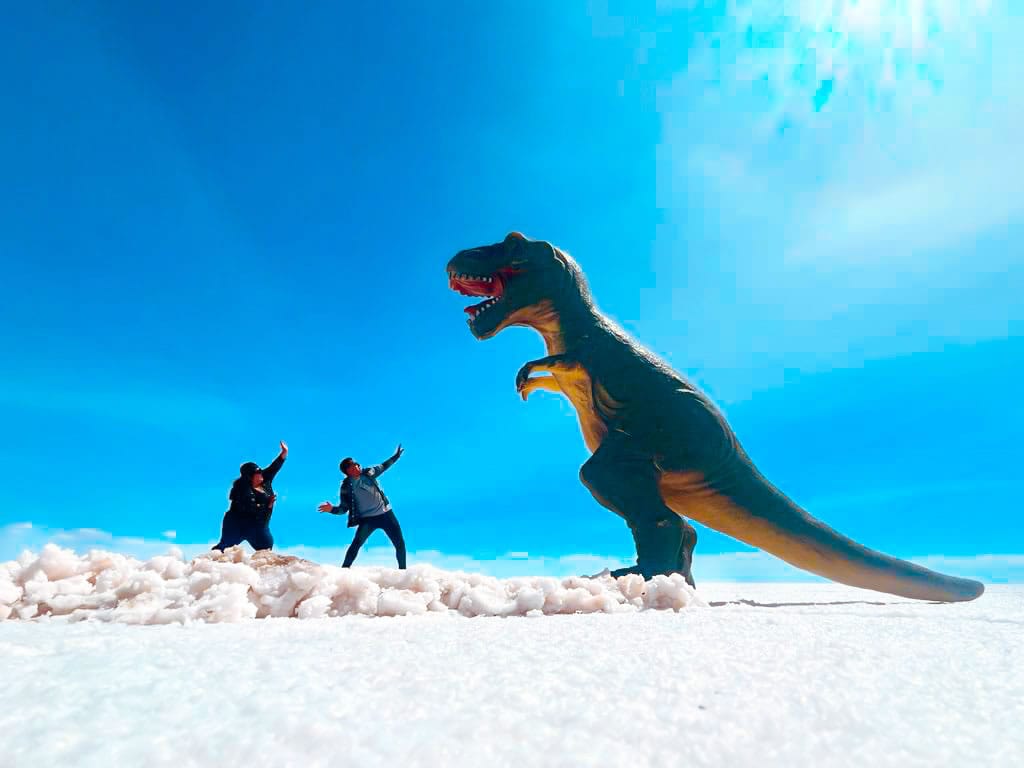 Escena de perspectiva forzada en el Salar de Uyuni: un dinosaurio de juguete gigante parece asustar a dos aventureros, quienes reaccionan con sorpresa ante la ilusión de su inmensidad en el desierto de sal.