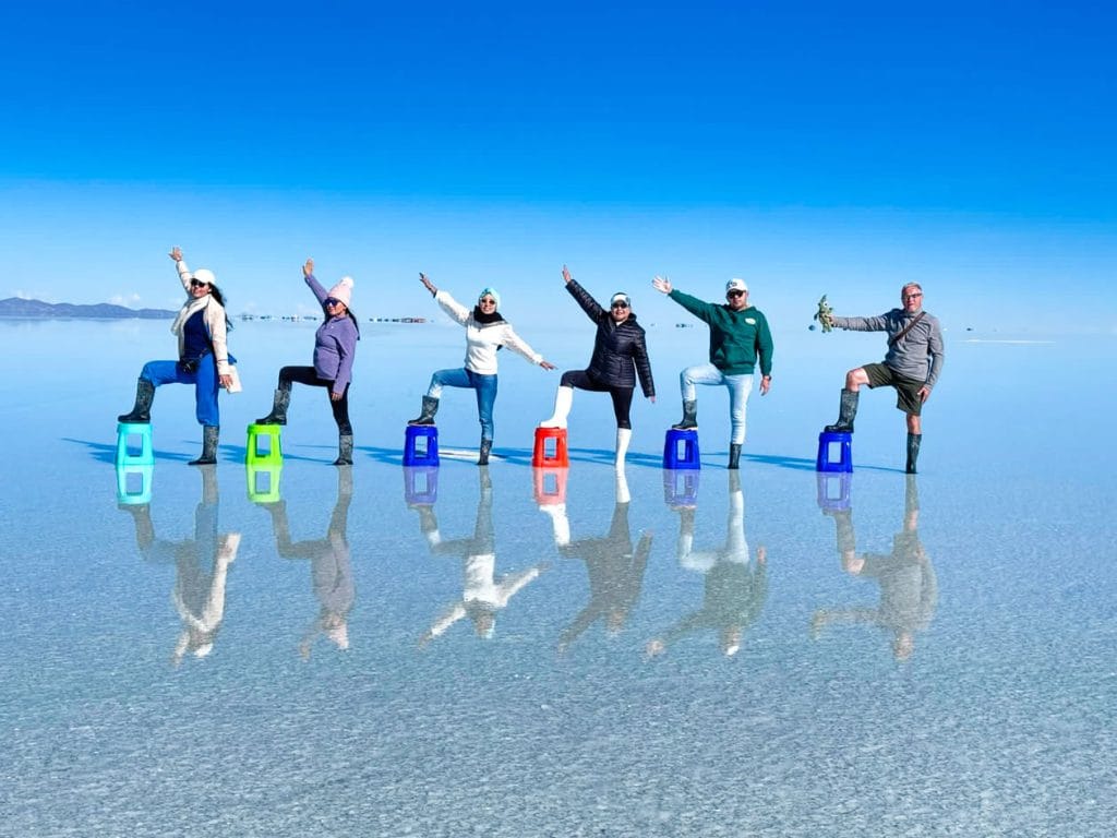 Grupo de viajeros posando creativamente en el Salar de Uyuni, apoyando un pie en el suelo y el otro en una banca de color, creando una divertida ilusión de perspectiva forzada en el horizonte blanco.