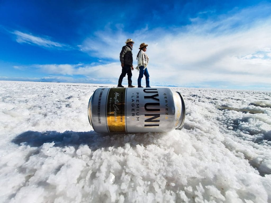 Ilusión de perspectiva forzada en el Salar de Uyuni: una pareja de viajeros simula caminar sobre una gigantesca lata de cerveza con el logo UYUNI, aprovechando el horizonte blanco para el efecto visual.