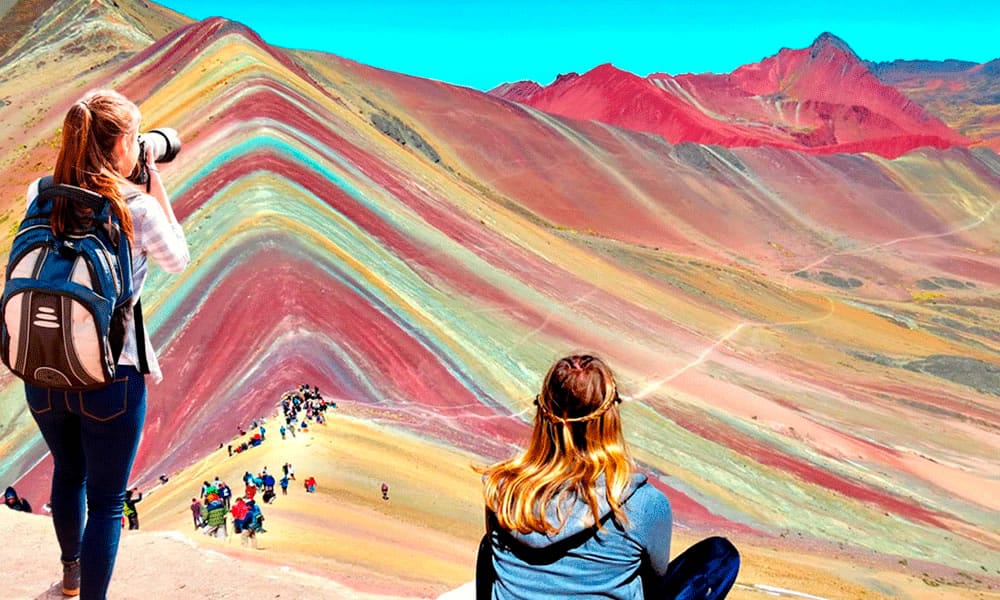 Dos turistas mujeres en primer plano, una tomando fotos y la otra contemplando el paisaje de los Andes peruanos en la caminata a la Montaña de Siete Colores (Vinicunca).