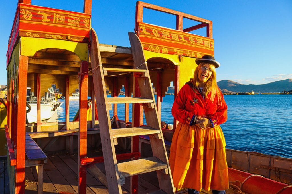 Turista alegre posando en lancha con vestimenta típica de los Uros, destacando la decoración con tejidos folclóricos de la embarcación en el Lago Titicaca.