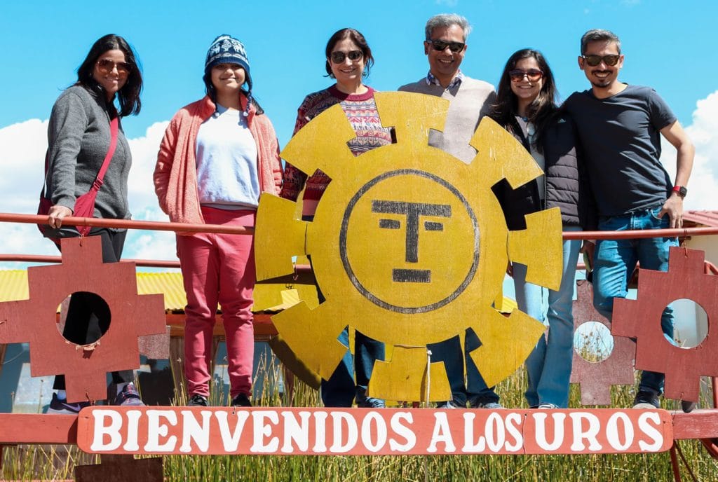 Turistas posando detrás del letrero de bienvenida a los Uros, destacando los símbolos andinos de Sol y Chacanas.