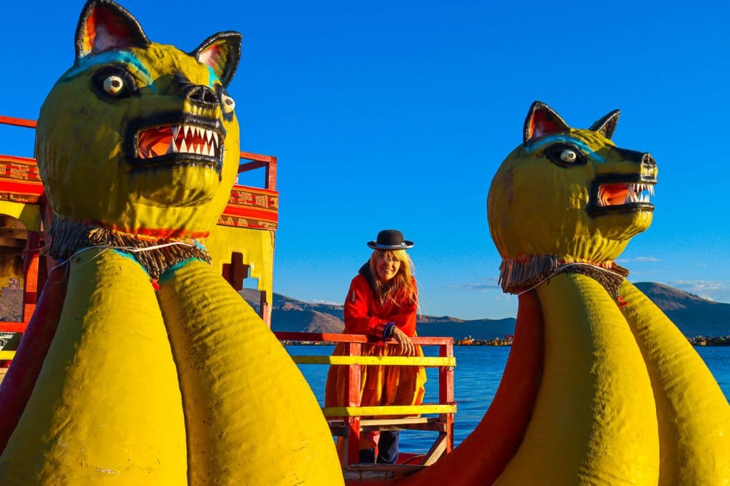 Balsa catamarán artesanal de totora navegando el Lago Titicaca, con proas decoradas en forma de cabezas de puma; turista con vestimenta típica a bordo de la misma.