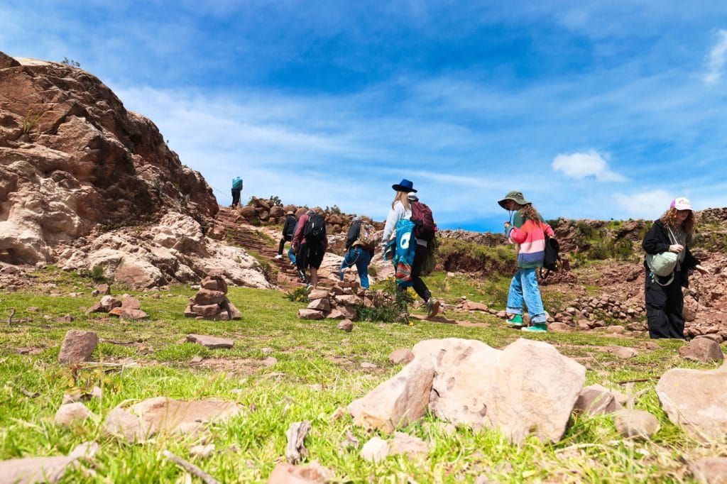 Turistas caminan por el sendero rústico de ascenso en la Isla Taquile, disfrutando de las vistas panorámicas del Lago Titicaca, Puno.